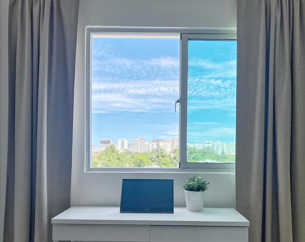 A window provides a view of the city skyline, featuring modern buildings and blue skies. Below, a small potted plant is placed on a white dresser, accented by light gray curtains that frame the window, creating a bright and airy environment.