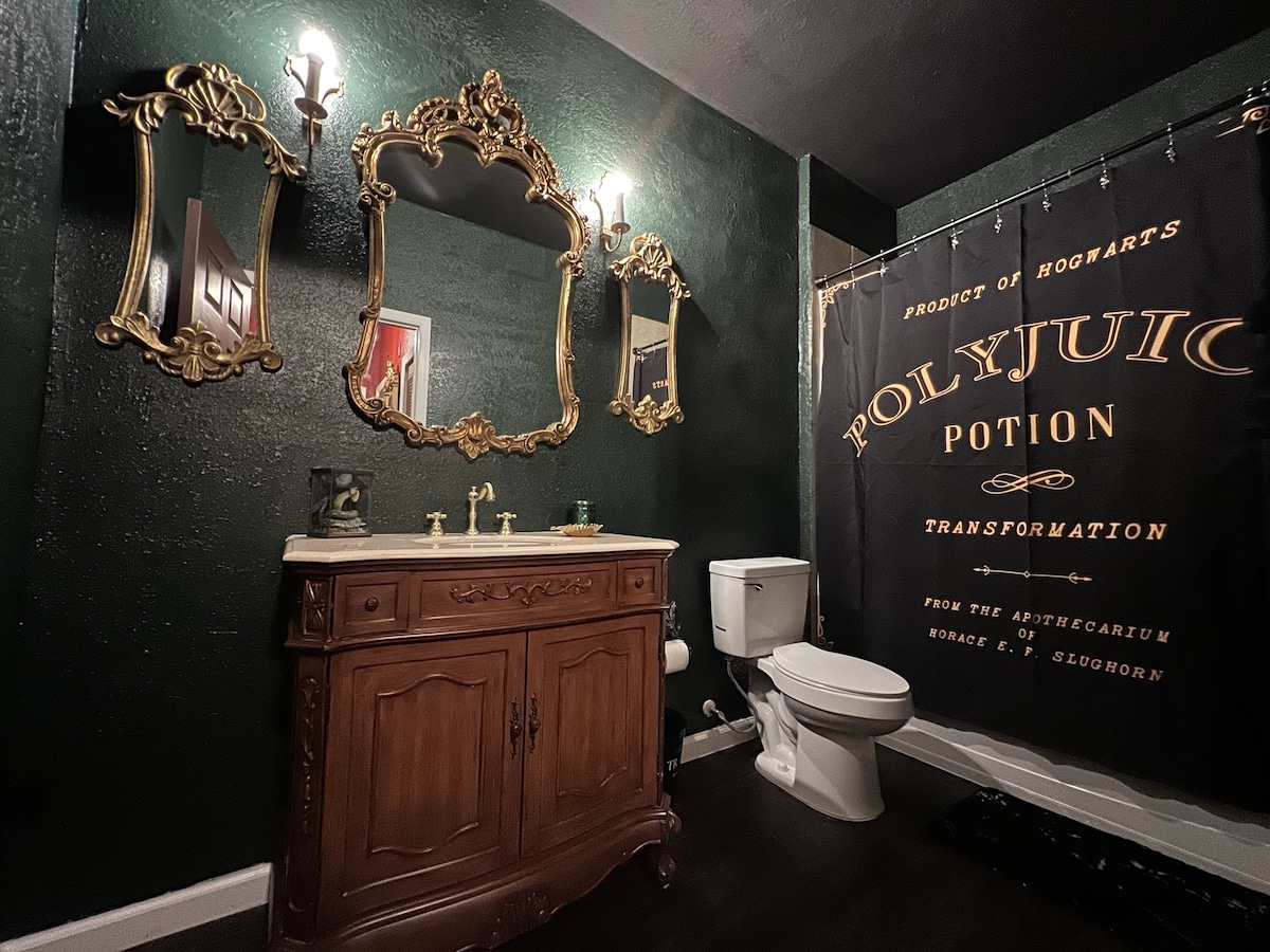 A dark green lavatory is adorned with gold-framed mirrors and a vintage-style wooden vanity, enhancing its classic aesthetic. A decorative shower curtain featuring 'Polyjuice Potion' adds a whimsical touch. The space is equipped with a standard toilet and ample lighting from wall sconces.