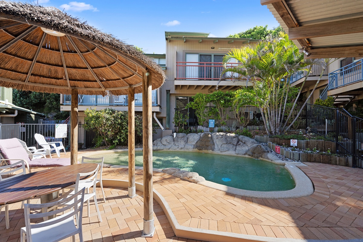 An outdoor area features a thatched gazebo with a wooden table and chairs beside a curved swimming pool. Lush greenery surrounds the pool, and a rocky landscape can be seen along one edge. The two-story building is located in the background.