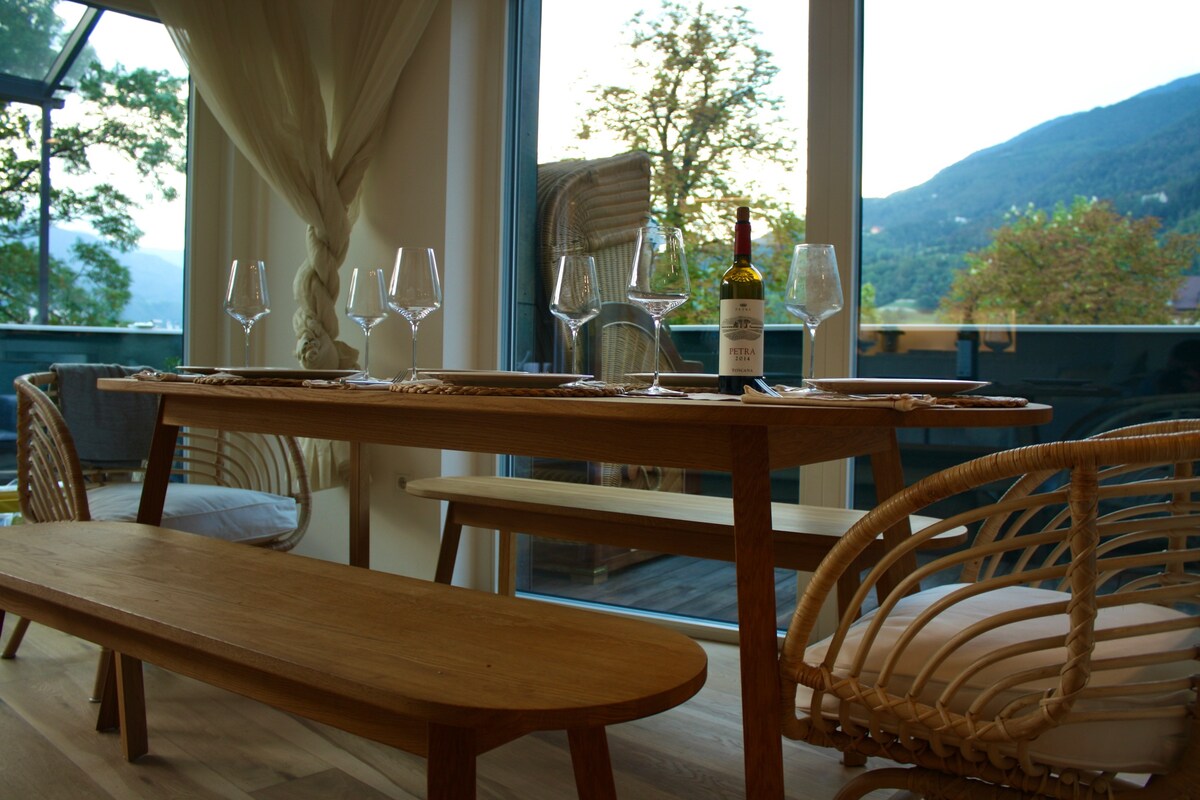 A natural wood dining table is set with wine glasses and a bottle of wine, creating a welcoming dining area. Rattan chairs surround the table, and large glass doors provide a view of the mountains and outdoor space, allowing for ample natural light.