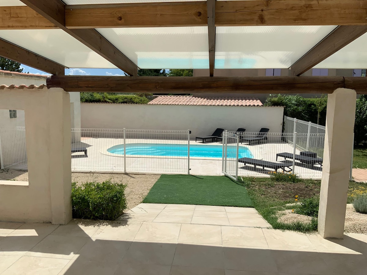 A private pool area is enclosed with a secure fence. The clear blue water is complemented by several lounge chairs arranged neatly around the pool. A shaded terrace with wooden beams provides a comfortable view of the outdoor space, enhancing the tranquility of the setting.