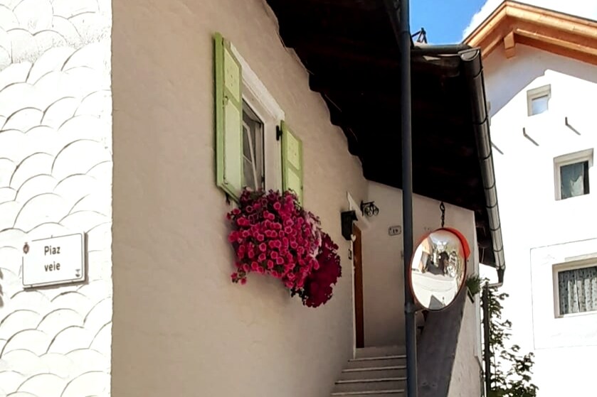 A charming exterior features a pale wall adorned with vibrant pink flowers cascading from a window frame with green shutters. A round mirror reflects the surrounding environment, enhancing the welcoming atmosphere of the entrance, which is flanked by stairs leading up to the home.