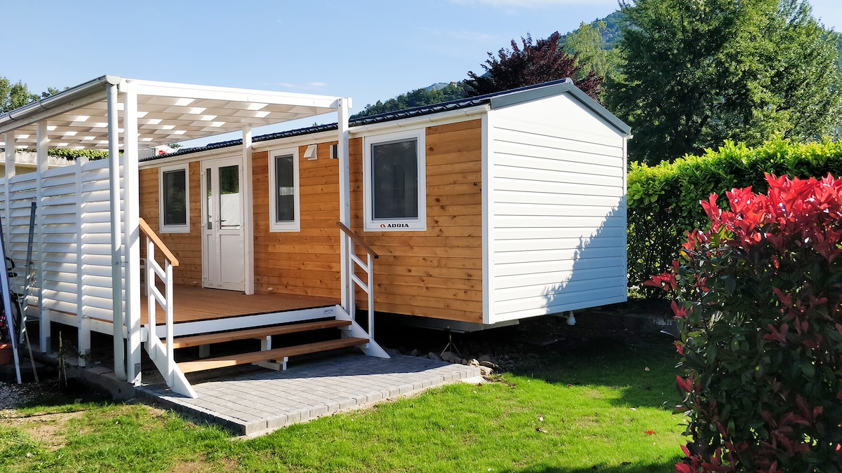 A modern mobile home is presented with a wooden exterior, featuring a large deck accessed by white steps. Surrounded by greenery, the structure features multiple windows, while vibrant plants in the foreground add color. The area is complemented by a well-maintained lawn.