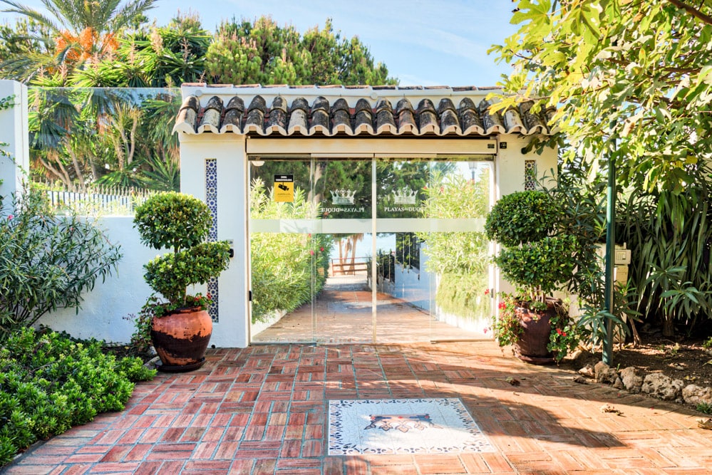 A welcoming entrance is lined with greenery, featuring a pathway of terracotta tiles. A decorative tile art piece is set into the ground, complemented by large potted plants on either side. Glass doors provide a view into the property, inviting guests to enter.