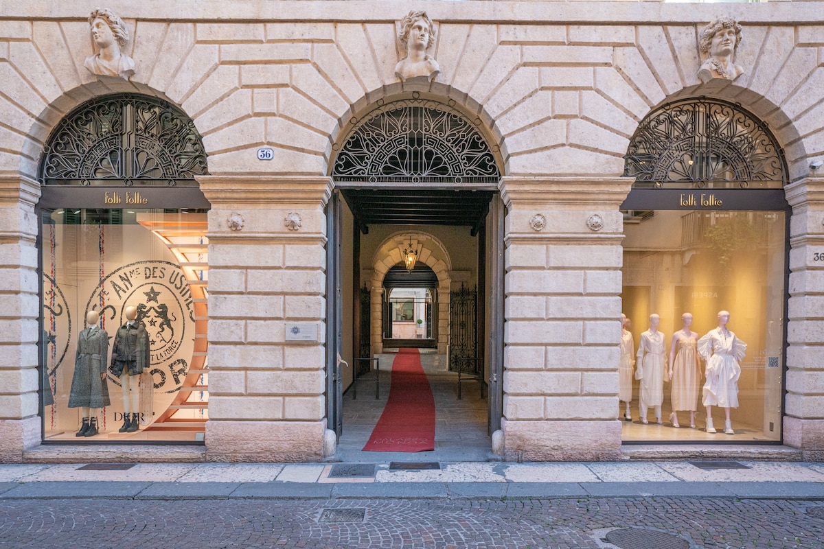 The image features an elegant entrance to a historic building, marked by arched doorways and intricate wrought-iron detailing. A red carpet leads through the arches, flanked by large shop windows displaying stylish apparel. The soft lighting highlights the architectural beauty and inviting ambiance.