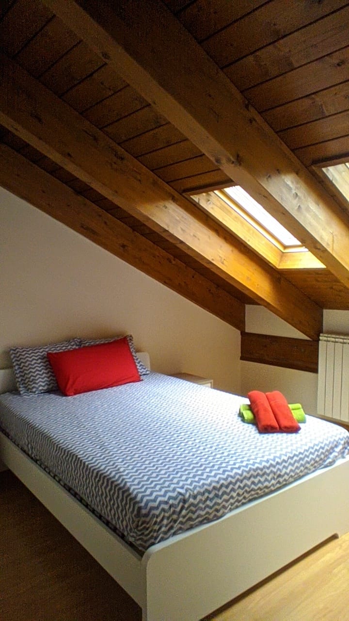 A cozy bedroom features a spacious bed with patterned bedding and bright accent pillows. Natural light enters through skylights, illuminating the wooden ceiling beams. Towels are neatly arranged at the foot of the bed, adding a pop of color to the serene space.