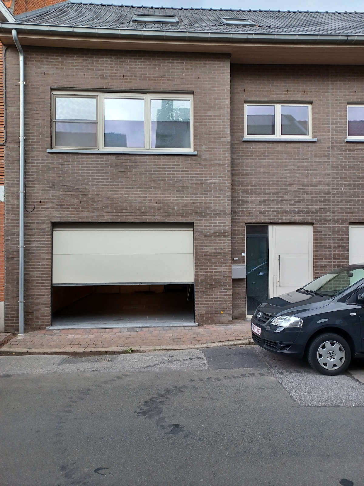 The image shows a modern, two-story building featuring a brick exterior. A garage door is visible on the left, slightly open, revealing a portion of the interior. The right side displays a door leading to the entrance, framed by large windows above, allowing natural light.