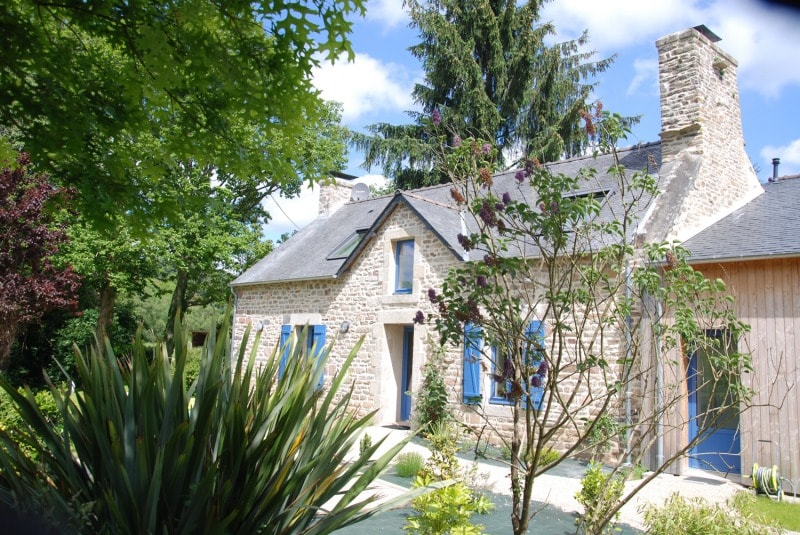 The charming stone cottage features blue window shutters and a slate roof. Lush greenery surrounds the home, with a mix of flowering plants and tall trees visible. A pathway leads to the entrance, providing a welcoming atmosphere.