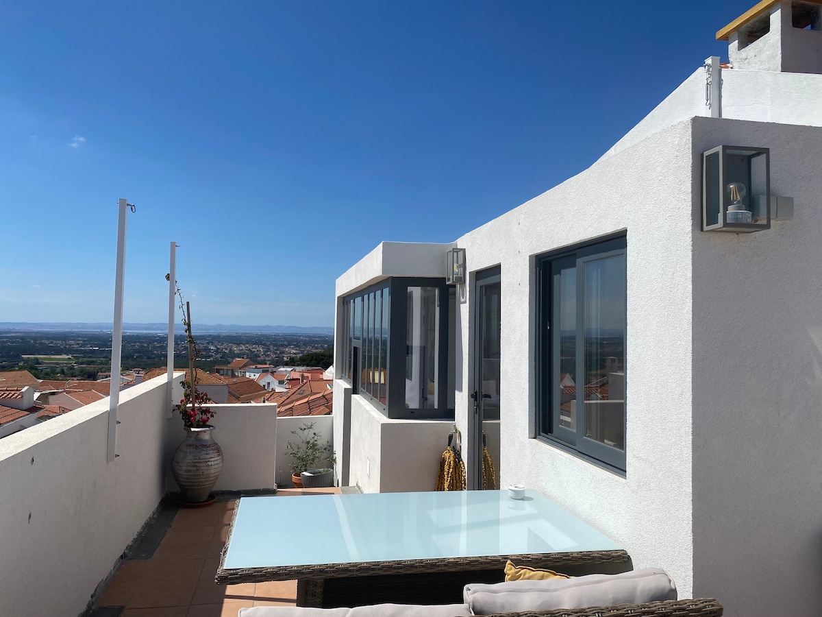 A terrace is featured with a clear glass table surrounded by comfortable seating, offering expansive views of the surrounding landscape. The white walls create a bright atmosphere, while a decorative plant adds a touch of greenery. The horizon stretches towards the ocean under a blue sky.
