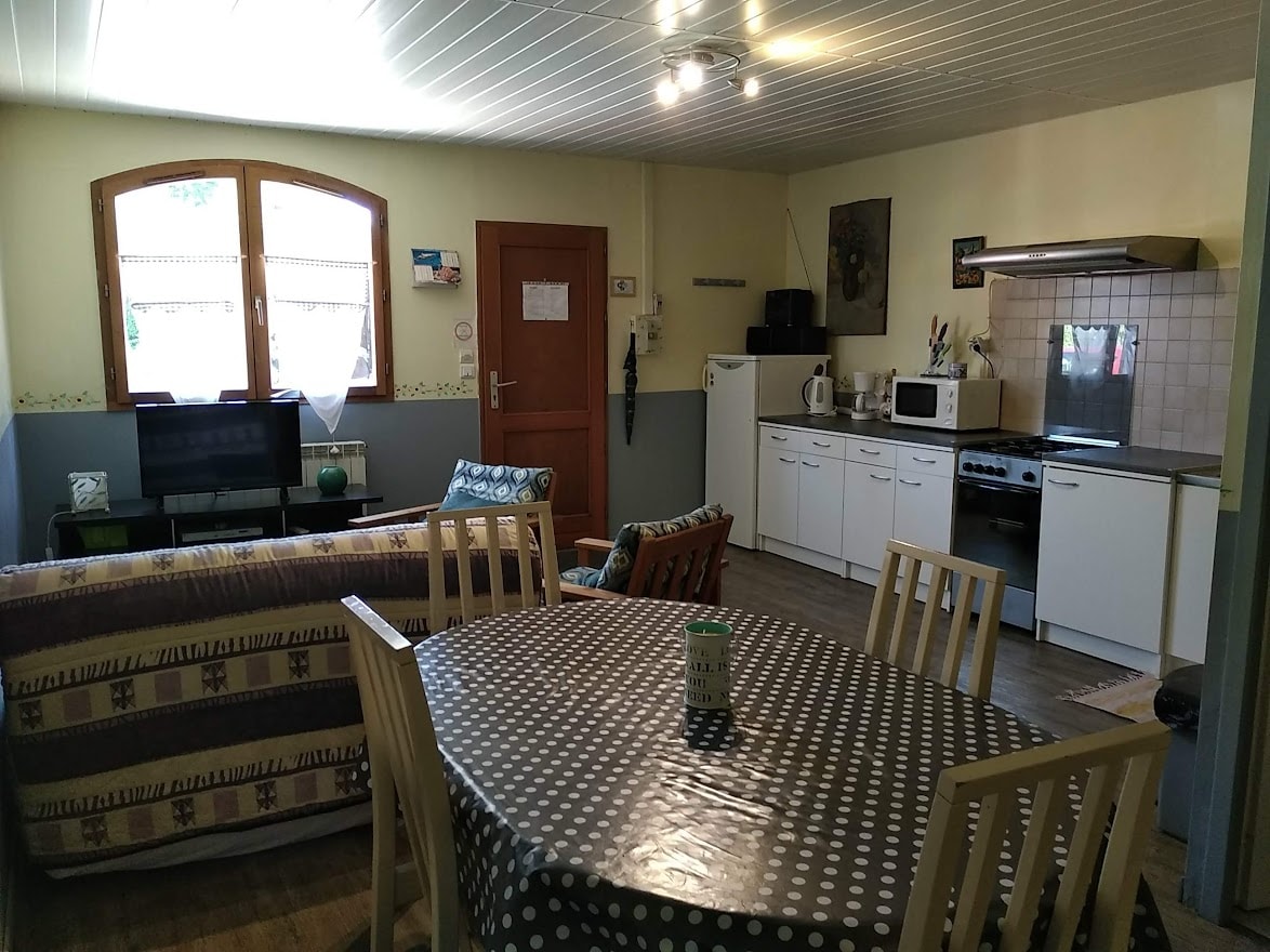 A bright living area features a dining table surrounded by several wooden chairs. A television is positioned across from a comfortable seating arrangement. The kitchen, equipped with modern appliances, is visible along one wall. Natural light pours in through two arched windows.