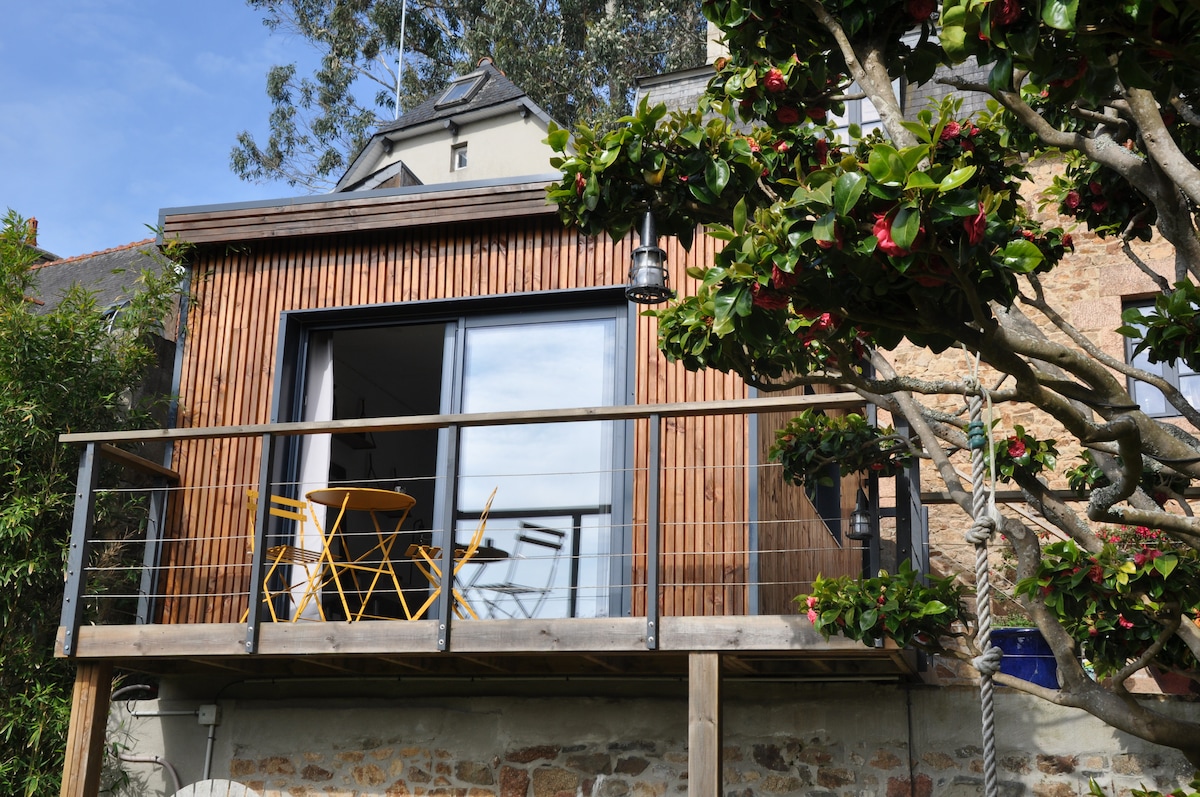 A wooden terrace overlooks a garden, featuring two yellow chairs and a small table. Large glass doors provide natural light to the interior space, framed by greenery from nearby plants and trees, creating a serene outdoor area.