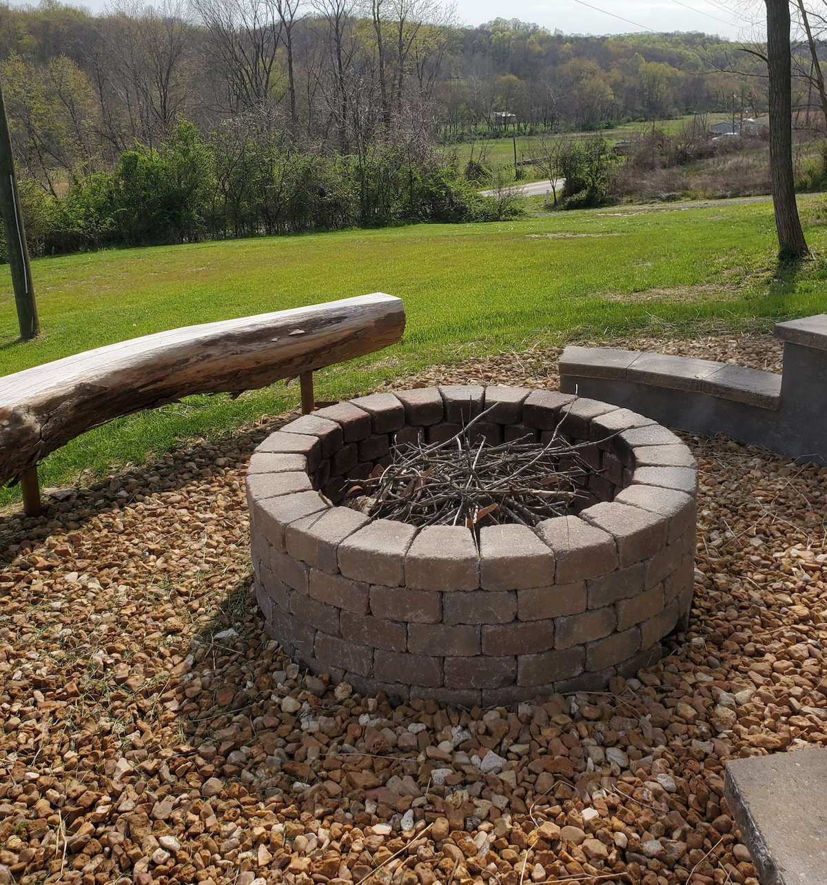 A stone fire pit is centrally located, surrounded by a gravel area. Natural logs serve as seating, providing a rustic touch. Lush green grass stretches out in the background, while trees and a peaceful landscape create a serene setting for outdoor gatherings.