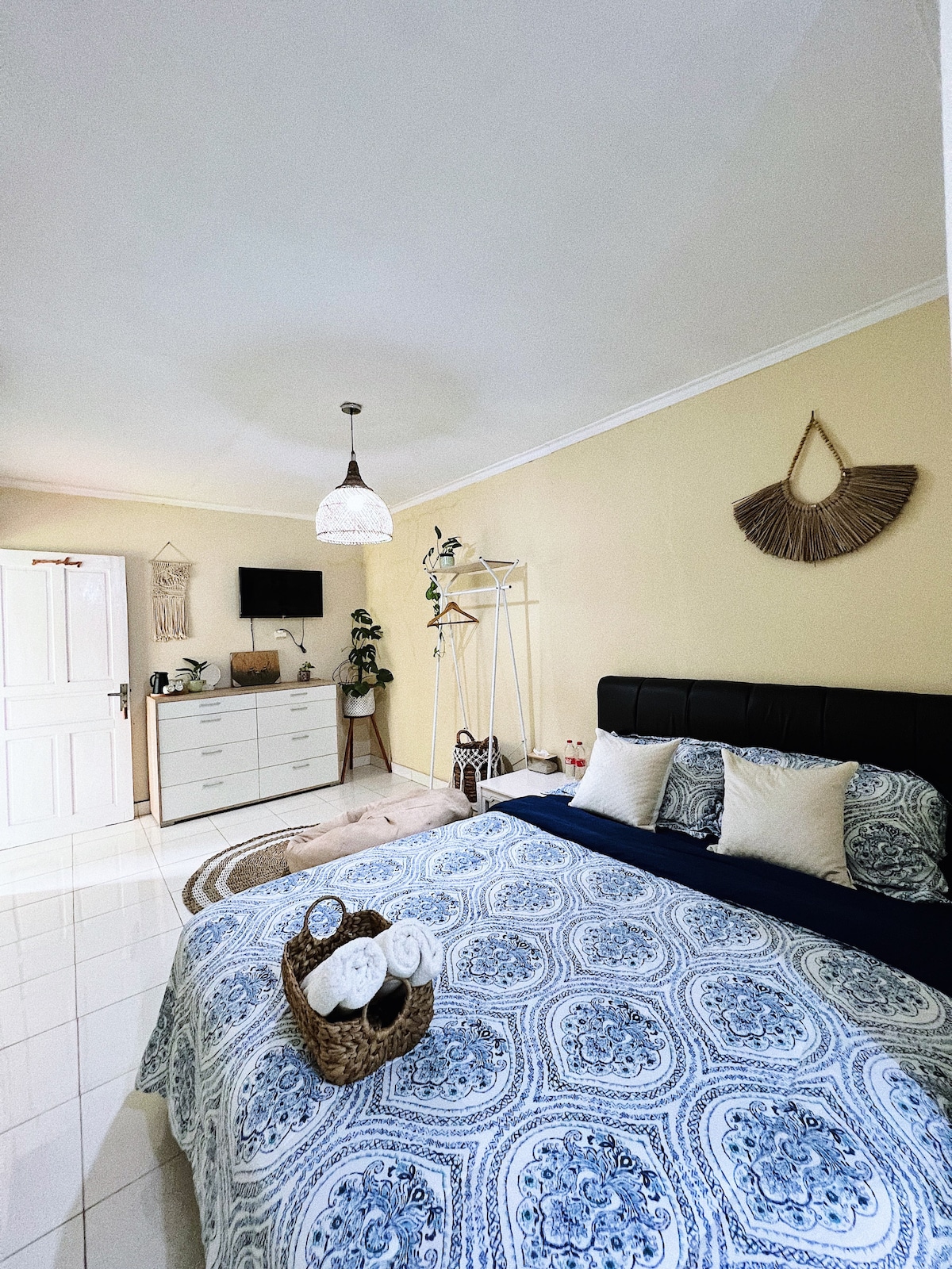 A well-lit bedroom features a spacious bed adorned with patterned bedding and decorative pillows. A stylish light fixture hangs overhead, while a dresser and decorative accents enhance the serene ambiance. A television is mounted on the wall, contributing to the room's convenience.