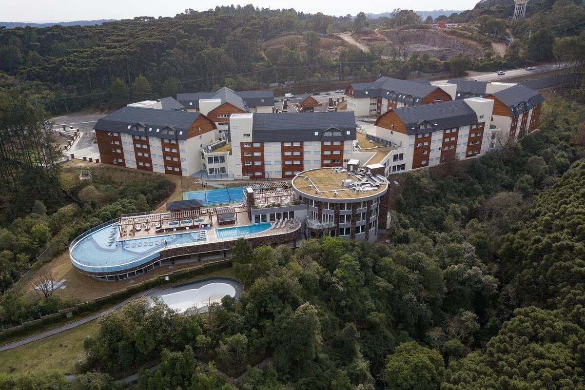 Aerial view of a resort nestled within lush greenery, featuring multiple buildings with modern architecture. The complex includes pools and recreational areas, surrounded by trees. The layout emphasizes the harmony between the structures and the natural landscape.