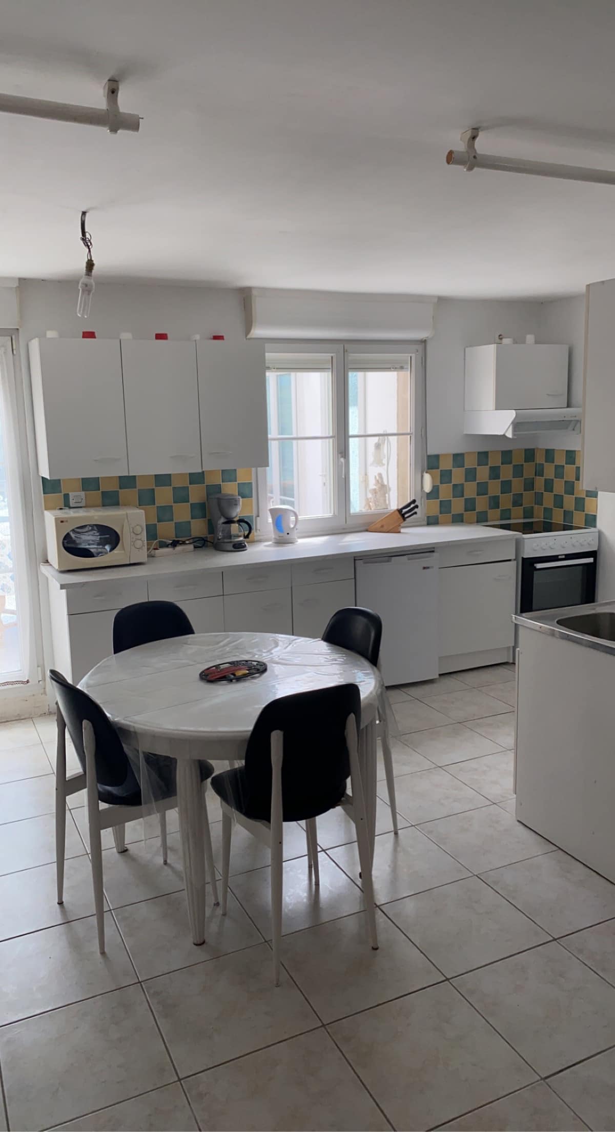 A functional kitchen features white cabinetry and a tiled backsplash in blue and yellow. A round table with four black chairs occupies the center of the space, while appliances including a microwave and a stove are neatly arranged. Natural light enters through the window.