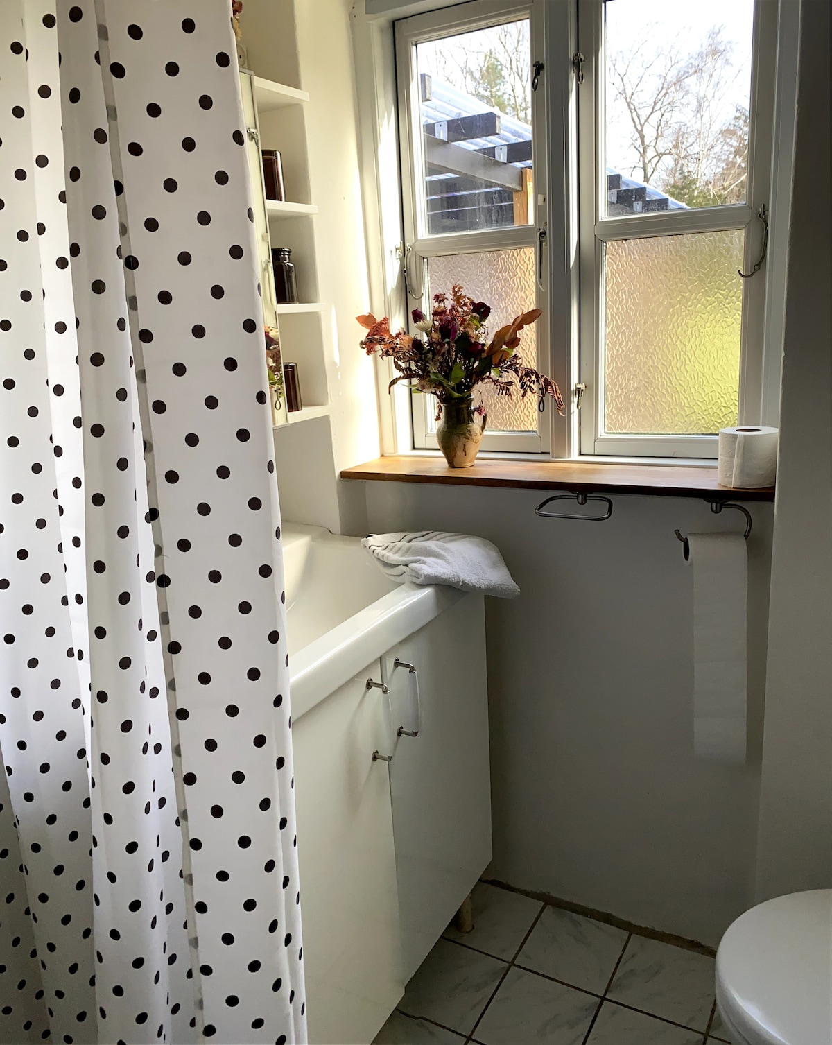A bathroom is shown, featuring a white sink and a toilet positioned beside a window. A floral arrangement sits on the windowsill, with natural light illuminating the space. A shower curtain with black polka dots adds a playful touch, and a towel is neatly placed nearby.