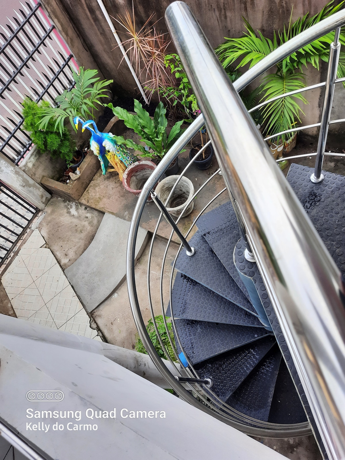 A spiral staircase is shown, featuring silver railings that contrast with a textured black surface. Below, a small outdoor space is visible, showcasing potted plants and a decorative statue. The area is surrounded by a concrete wall, offering a glimpse of greenery.