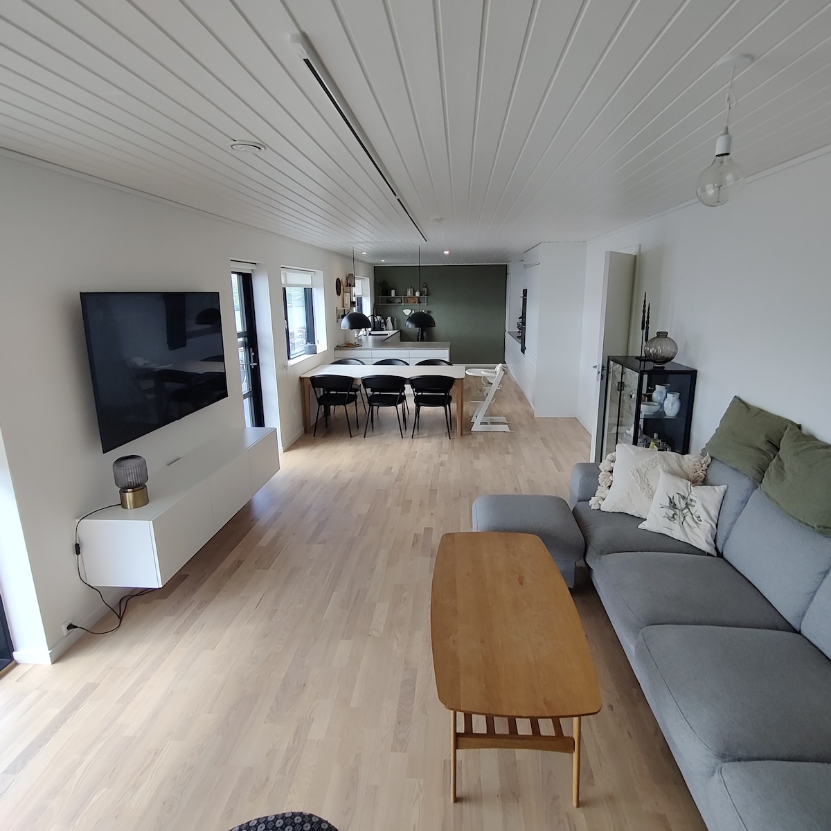 A light-filled living area features a comfortable gray sofa paired with a wooden coffee table. A flat-screen television is mounted on the wall opposite the seating. The dining area, visible in the background, is furnished with a long table and black chairs, fostering a welcoming space.