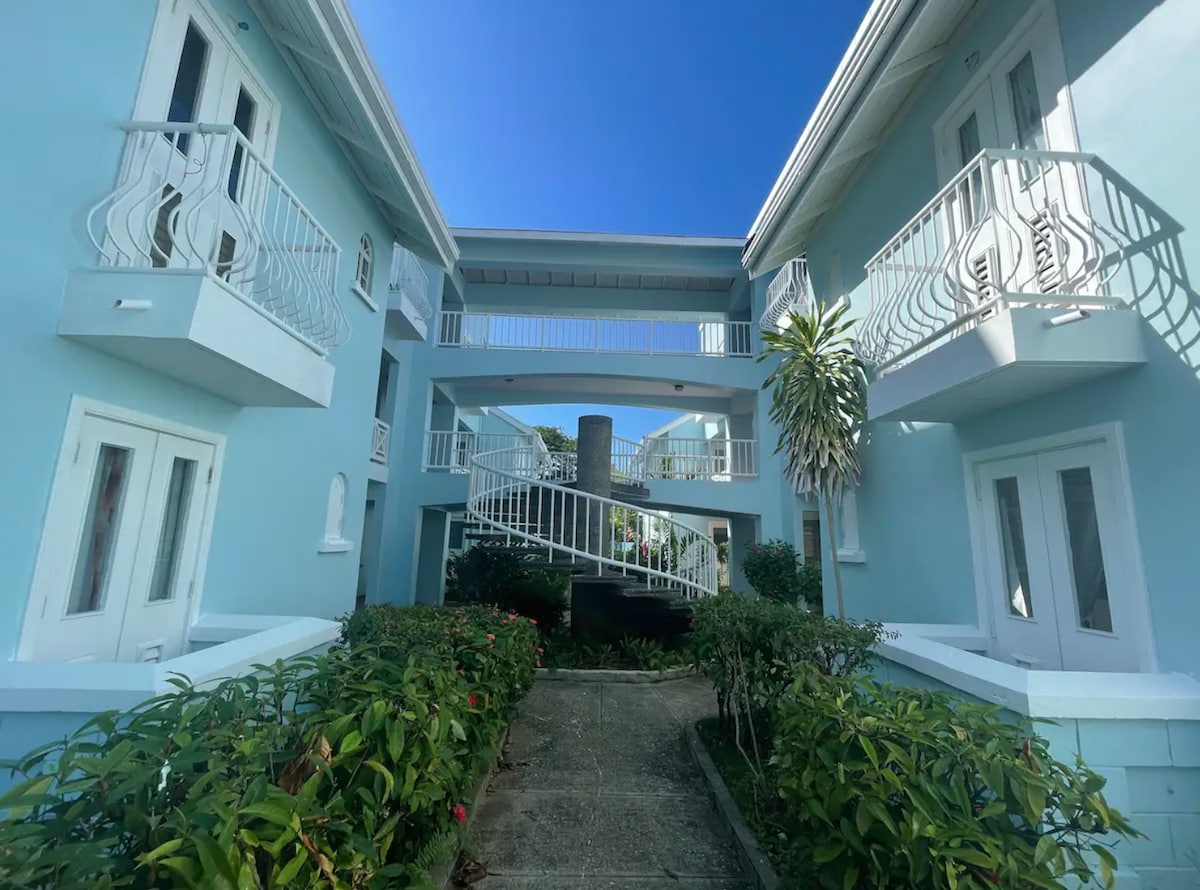 A light blue building is surrounded by well-kept greenery, featuring multiple balconies with decorative railings. A central pathway leads to a circular area with a small fountain, providing a sense of calm in the entryway.