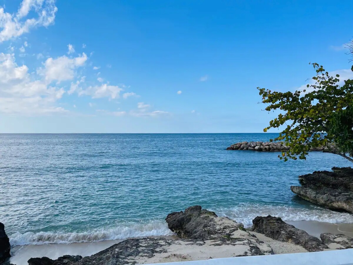 A serene view of the ocean stretches to the horizon, framed by soft waves lapping against rocky outcrops. A clear blue sky is dotted with a few white clouds, while gentle ripples reflect the sunlight on the water’s surface.
