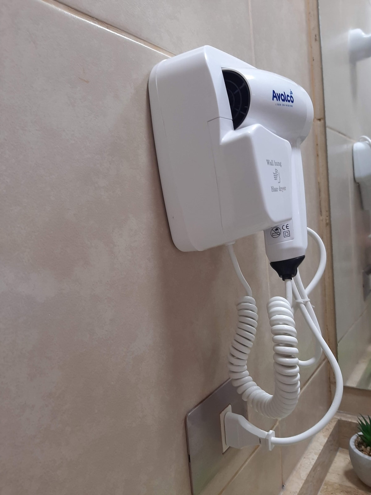 A wall-mounted hairdryer is displayed in the bathroom. The device is white and features a coiled cord for convenient use. The surrounding area includes neutral-colored tiles, contributing to a clean and functional bathroom space.