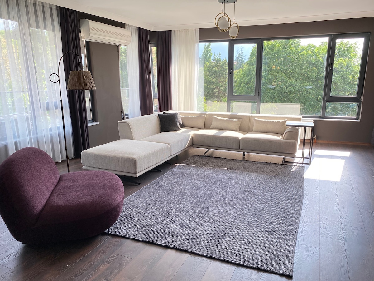 A spacious living area features a modern sectional sofa, a purple chair, and a large area rug. Ample natural light floods the space through floor-to-ceiling windows, which offer views of lush greenery outside. Simple decor provides a clean and open atmosphere.