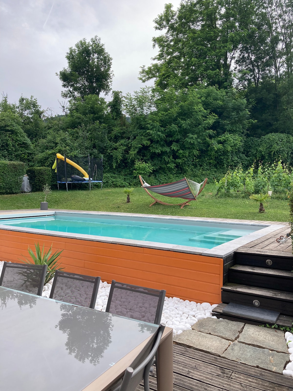 An outdoor area features a swimming pool surrounded by greenery. A colorful hammock hangs nearby, next to a trampoline and slide set. A dining table with several chairs is positioned on a wooden deck, accompanied by neatly arranged pebbles and plants.