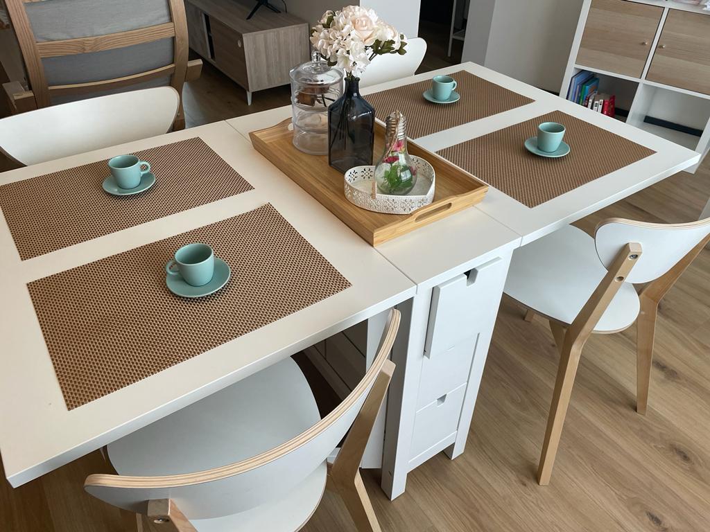 A dining area is presented with a white table featuring four placemats and light blue cups arranged neatly. A tray holding decorative items sits at the center. Chairs with light wooden frames are positioned around the table, enhancing the space's open feel.