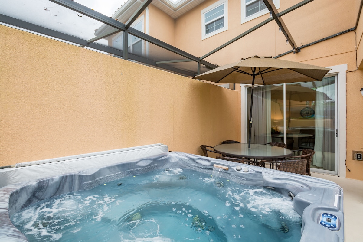 A private screened-in patio showcases a hot tub surrounded by a light orange wall. A large umbrella provides shade over an outdoor dining table with wicker chairs. Windows offer views of the surrounding area, enhancing the sense of openness.