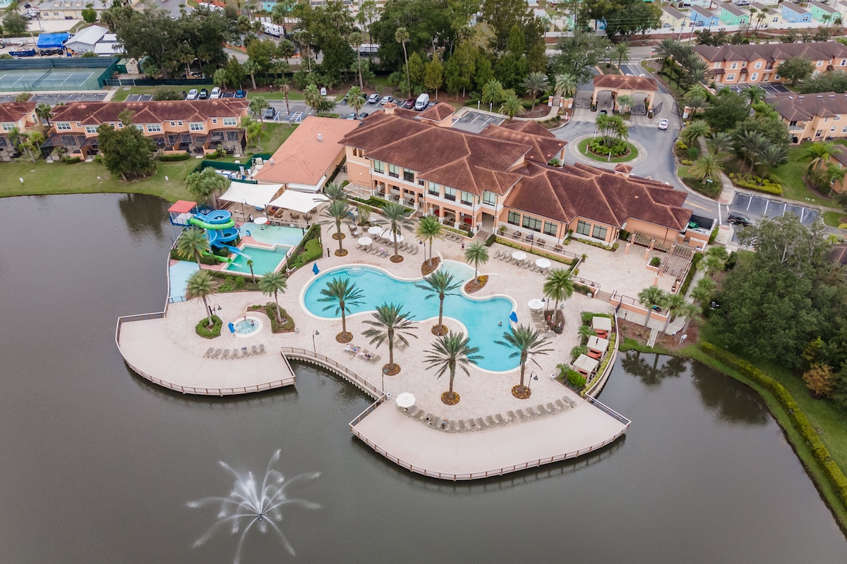 An aerial view of a resort featuring a large swimming pool surrounded by lounge chairs and palm trees. A water slide and children's play area are seen alongside a reflective lake with a fountain in the foreground, showcasing green landscapes and the adjacent townhomes.
