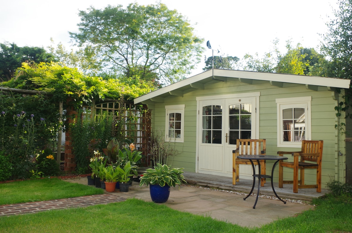 A charming garden space is presented with a light green cabin, featuring large windows and a door. A small seating area is positioned outside with two wooden chairs and a table. Lush greenery surrounds the cabin, enhancing the serene setting.