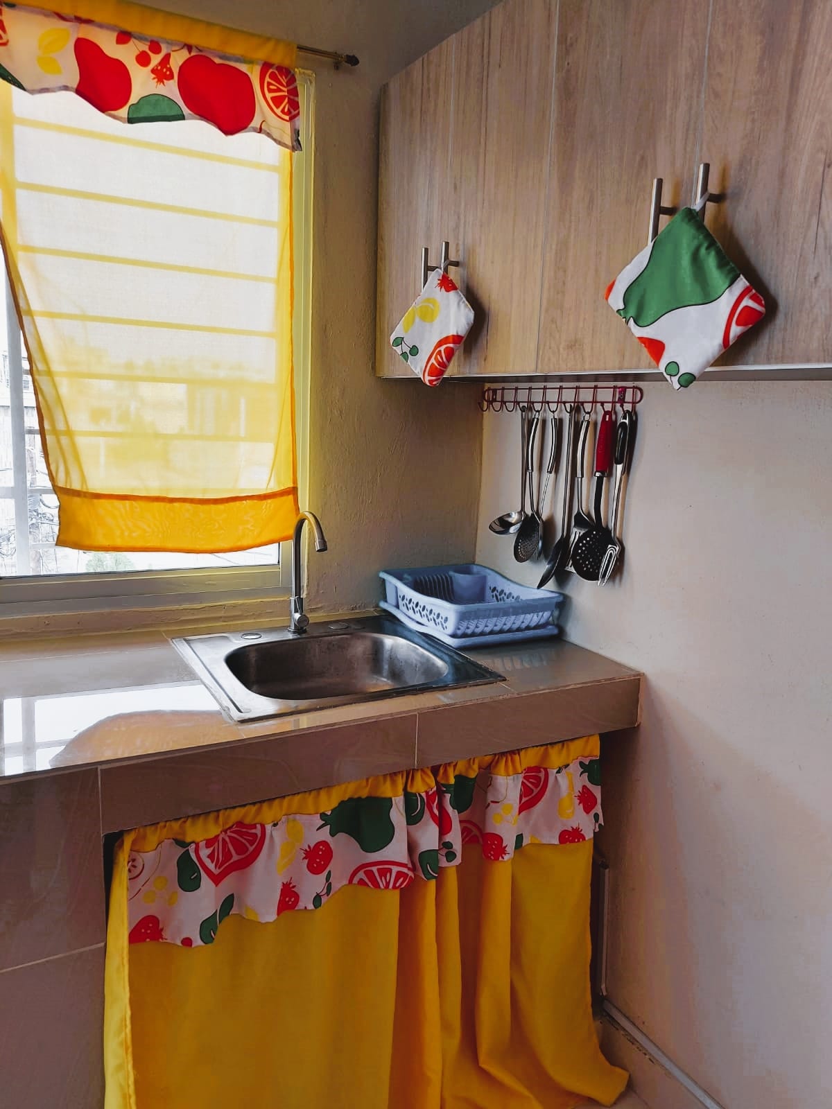 A well-organized kitchen area features a compact sink under a window adorned with a bright yellow curtain. Colorful dish towels and utensils are neatly arranged, while a fruit-themed curtain adds a cheerful touch to the space.