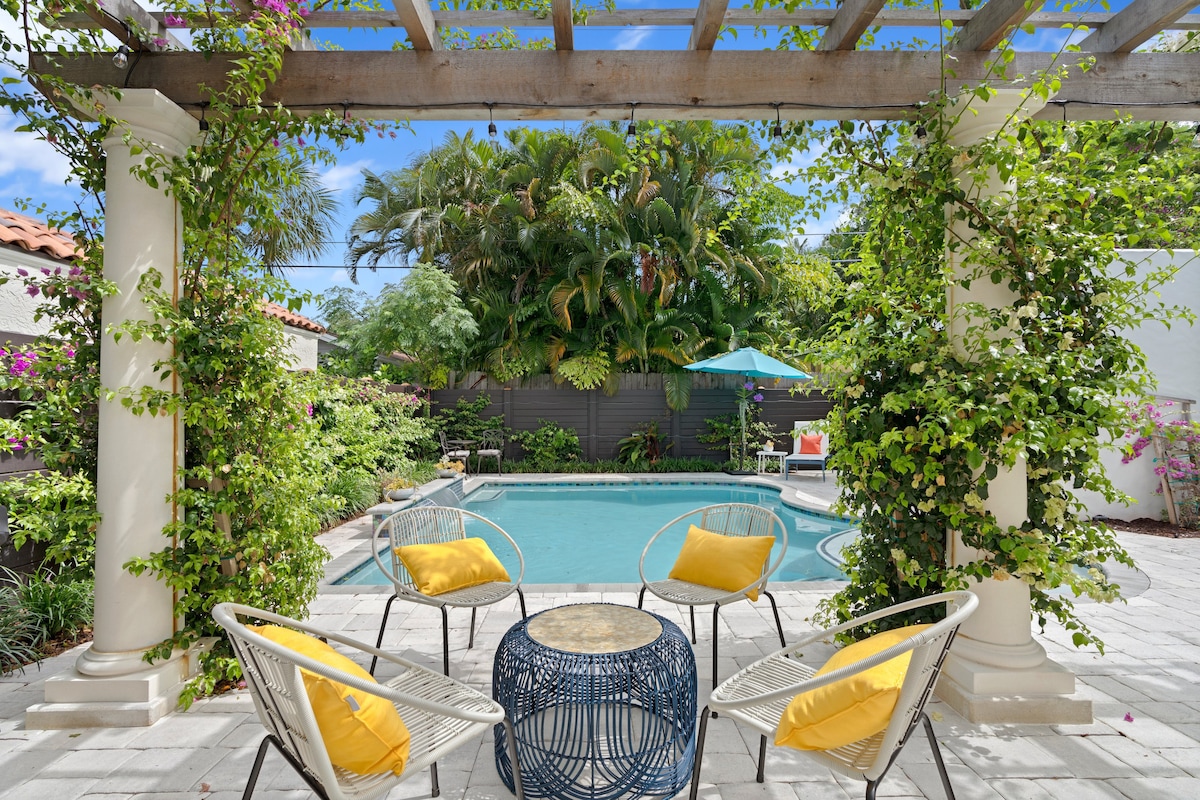 An outdoor living space is framed by a wooden pergola adorned with lush greenery. Four white chairs with yellow cushions surround a blue decorative table, offering a comfortable spot to relax. A turquoise sunshade and tropical plants are visible around the heated pool.