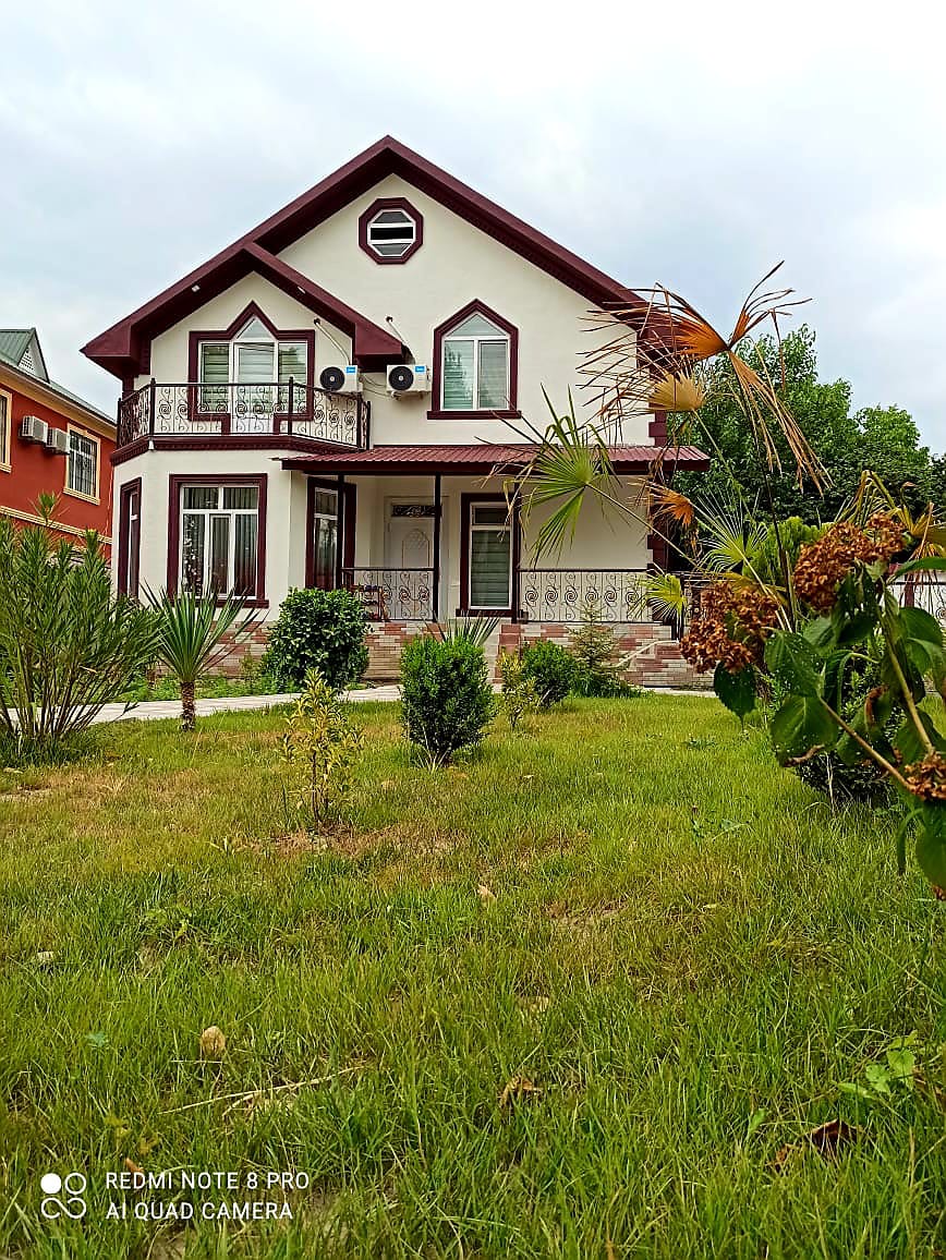 A two-story villa features a white facade accented with dark trim and a decorative balcony. Lush greenery surrounds the house, while a well-maintained yard offers a tranquil atmosphere. Multiple air conditioning units are visible on the exterior, providing comfort during warmer months.