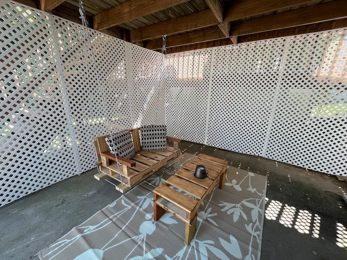 An outdoor seating area is presented with wooden furniture, including a low table and a swing-style couch. The space is enclosed by lattice panels, allowing light to filter through while providing privacy. A patterned rug lies on the concrete floor, enhancing the cozy atmosphere.