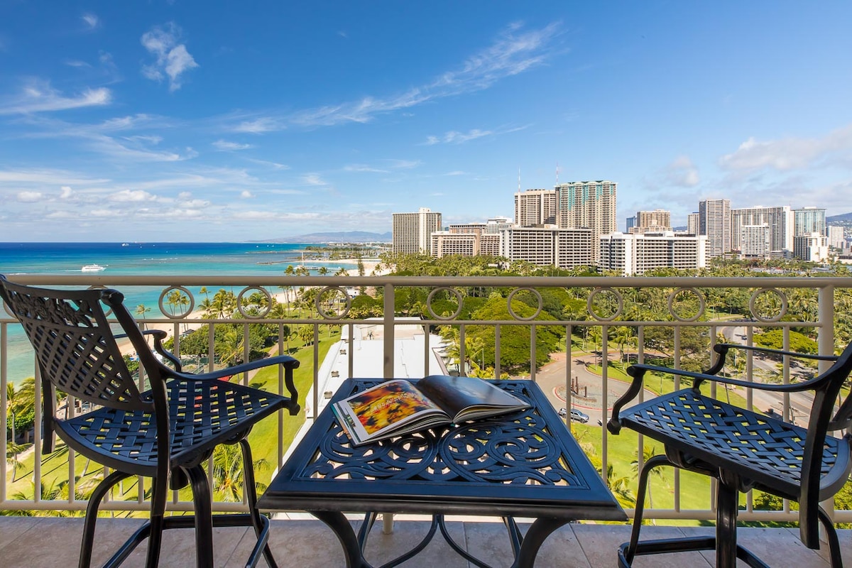 A balcony view presents a vibrant coastal scene, showcasing the ocean along with the Honolulu skyline. Two black metal chairs are positioned at a small round table, which is adorned with a magazine. Clear skies and bright sunlight enhance the inviting atmosphere.