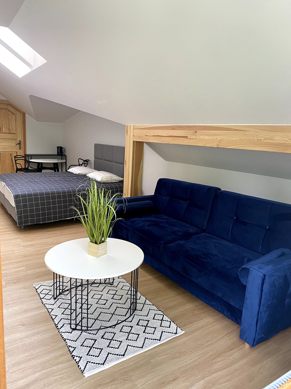 A cozy room is featured, showcasing a blue sofa near a small coffee table on a patterned rug. A bed with dark bedding is positioned in the background, and a plant in a gold pot adds a touch of greenery. Natural light enters through a skylight.