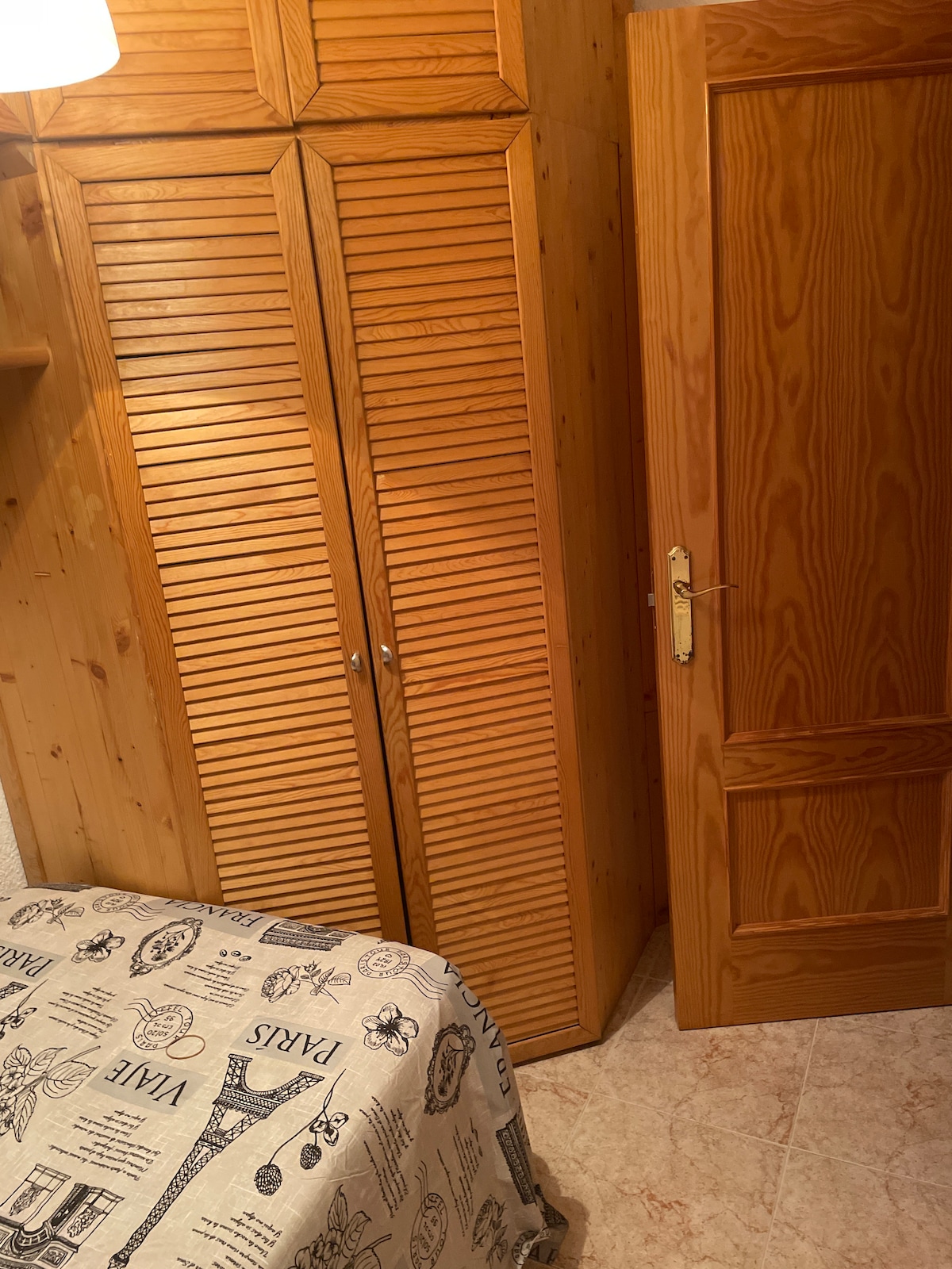 A wooden wardrobe with louvered doors is featured against a wall, providing ample storage space. Beside it, a complementary wooden door adds to the room's warm tone. A patterned bedspread can be seen draping over the bed, enhancing the overall simplicity of the cozy space.