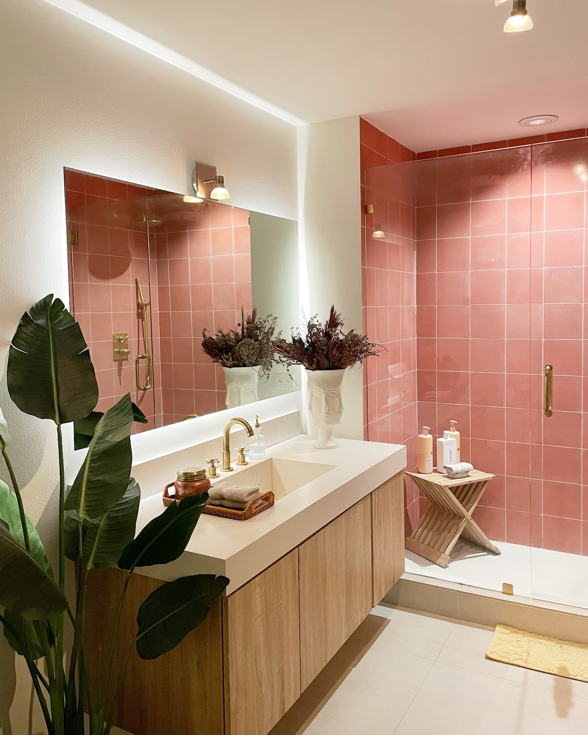 A modern bathroom features warm pink tiles, creating a gentle ambiance. A spacious vanity with a white countertop is complemented by a stylish mirror and elegant fixtures. Decorative plants and a geometric stool enhance the space, accompanied by various toiletries displayed thoughtfully.