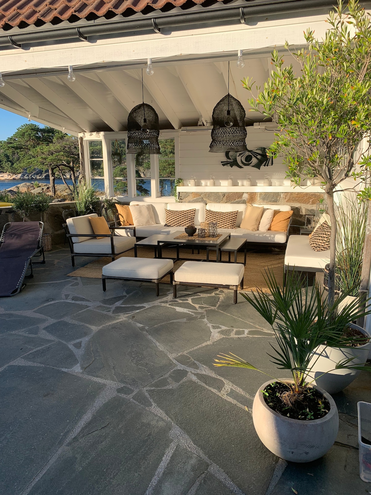 A welcoming outdoor seating area is showcased, featuring comfortable couches adorned with neutral-colored cushions. A table is centered among the seating, surrounded by decorative plants and stylish hanging light fixtures. The stone-paved floor adds a rustic touch to the inviting space.