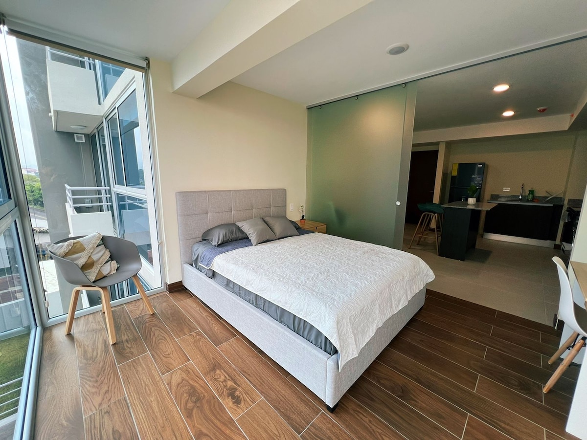 A bright sleeping area features a spacious bed with neutral bedding and gray accents. Natural light floods through large glass doors, providing views of the outdoor space. Modern furniture, including a chair and a small bedside table, complements the contemporary design.