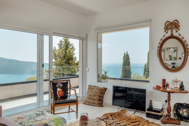 A stylish living room is presented with large glass doors allowing natural light and stunning views of the sea and hills. A decorative mirror enhances the space, while a mixture of seating arrangements includes a patterned chair and a cozy cushion. The room's decor reflects a modern aesthetic.