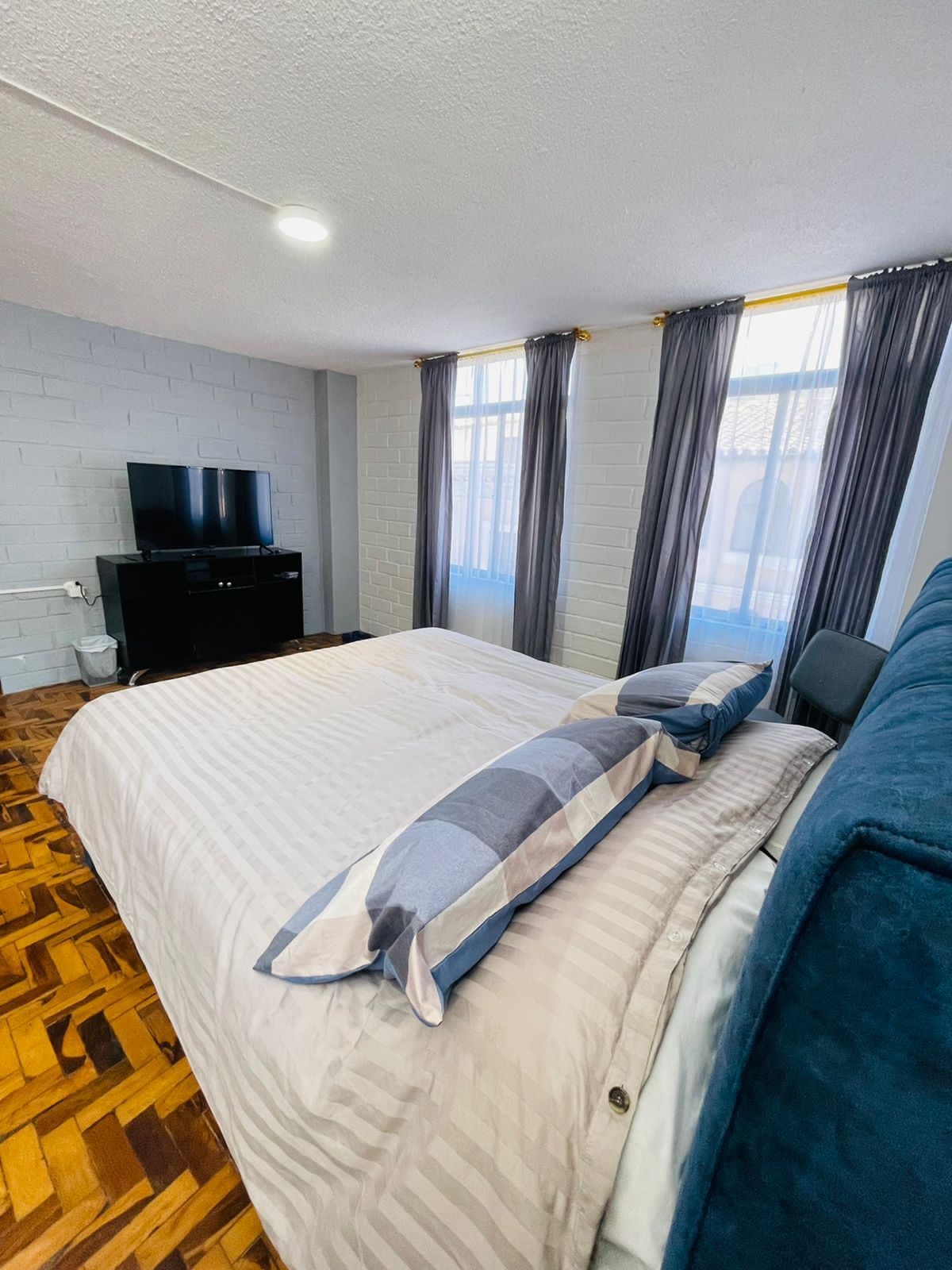 A spacious bedroom is presented with a large bed featuring soft linens and decorative pillows. A television is placed on a stand opposite the bed. Natural light filters through the tall windows, which are complemented by long, flowing curtains.