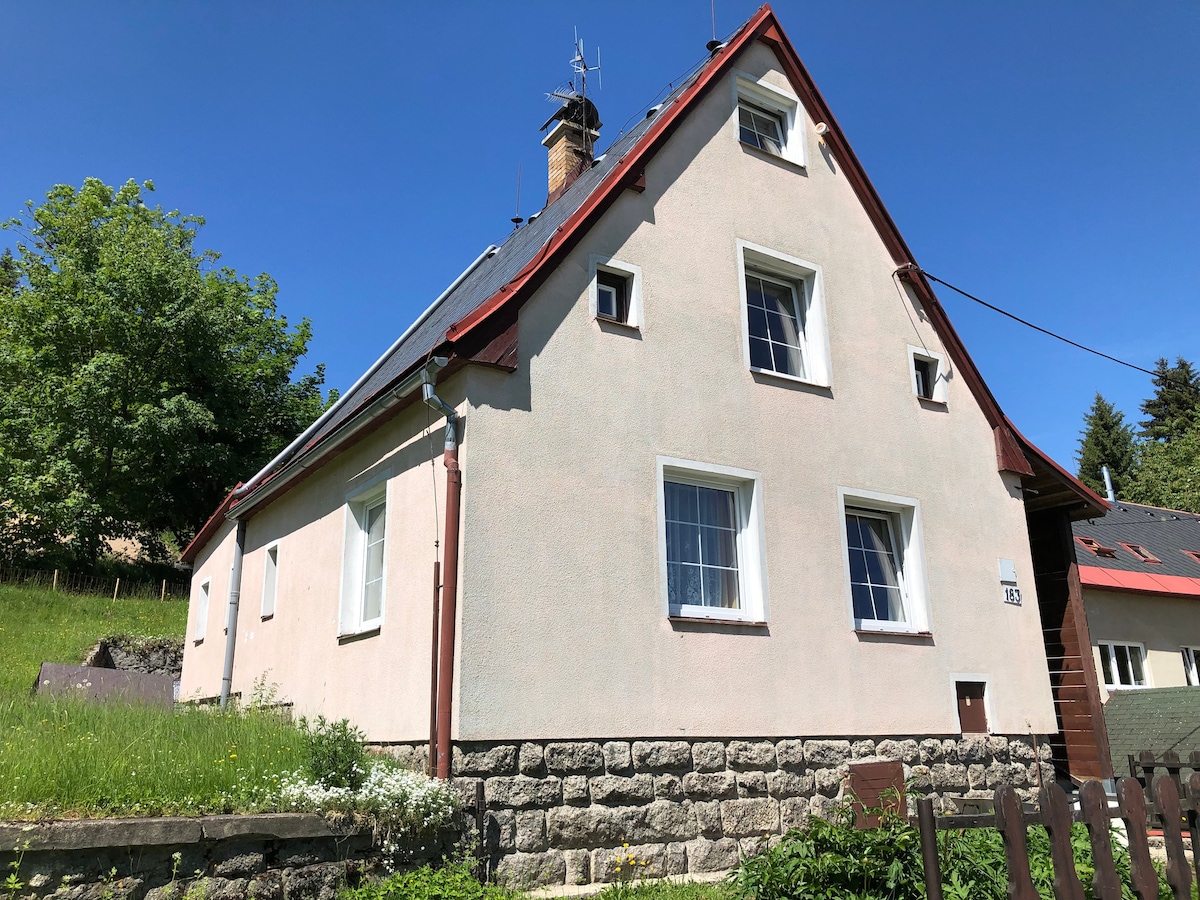 A charming two-story house is viewed from the side, featuring a light-colored exterior with three large windows on the upper level. The sheltered roof is complemented by greenery surrounding the property, while a stone wall forms the foundation along the base.