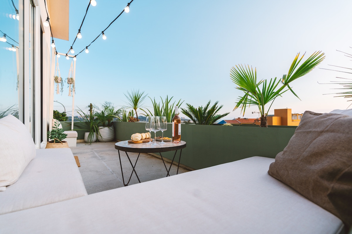 A spacious terrace features comfortable seating with soft cushions and a round table set for drinks. Decorative plants line the edges, contributing to a relaxed ambiance. Decorative string lights above enhance the outdoor space, creating a welcoming environment for gatherings.