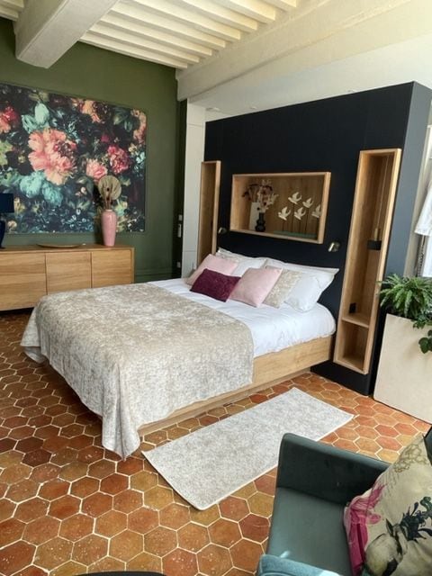 A spacious bedroom features a light-colored bed dressed in a textured blanket. Decorative pillows add soft hues against dark wood cabinetry. A large floral wall art piece brings color to the green accent wall, while hexagonal terracotta tiles cover the floor.