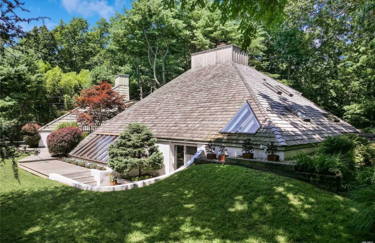 The contemporary home features a unique triangular roof with multiple skylights, surrounded by landscaped gardens. Mature trees provide natural shade, while smooth paths lead to the entrance. The overall design harmonizes with the lush green surroundings, creating a serene environment.