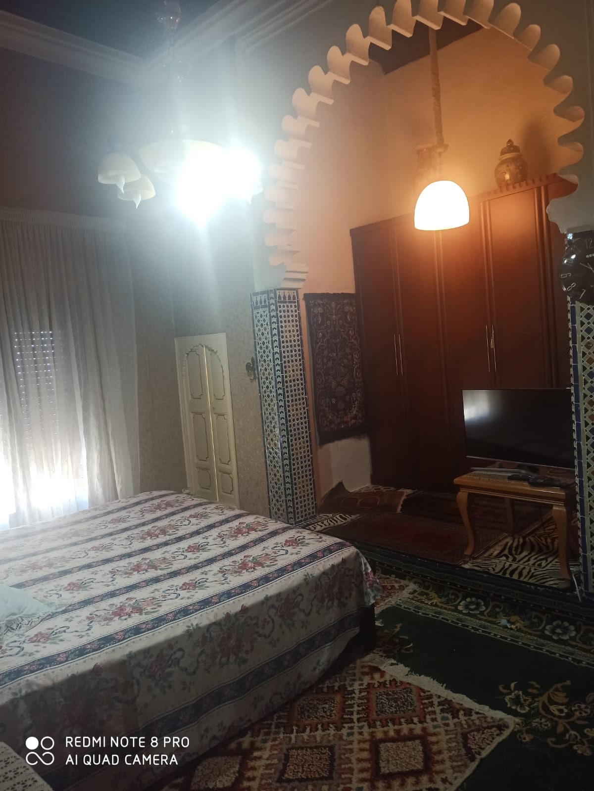 A bedroom features a spacious bed covered in a floral pattern. A wooden wardrobe stands against one wall, while a television is positioned nearby. Traditional rugs cover the floor, and natural light enters through tall windows adorned with sheer curtains.
