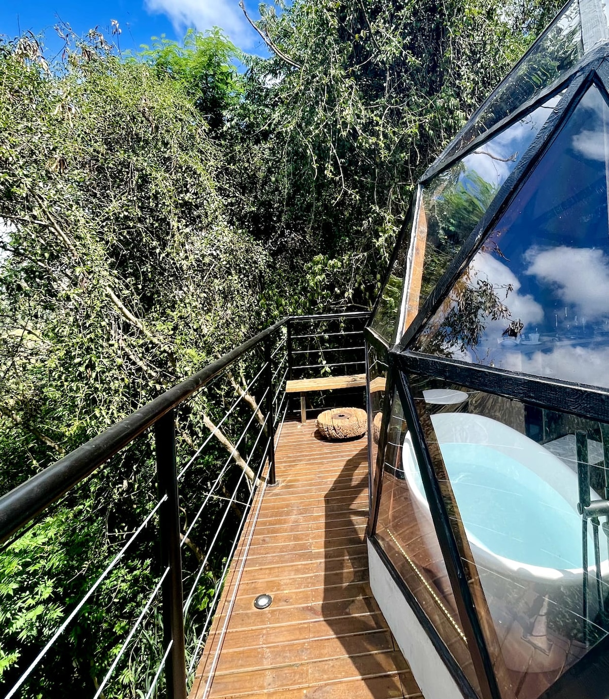 A wooden deck extends from the structure, overlooking lush greenery. A rounded bathtub can be seen through the large glass windows, inviting relaxation. A small bench is positioned beside a plush seating area, providing a tranquil space to enjoy the natural surroundings.