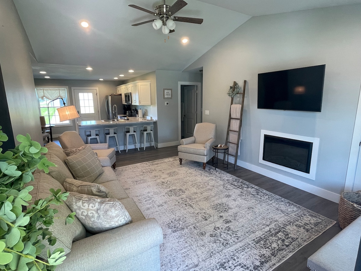 A light-filled living area features a comfortable sofa, a cozy armchair, and a stylish electric fireplace. A large wall-mounted television is visible above the fireplace, while a neutral-toned area rug anchors the space. The open layout connects seamlessly to the kitchen in the background.